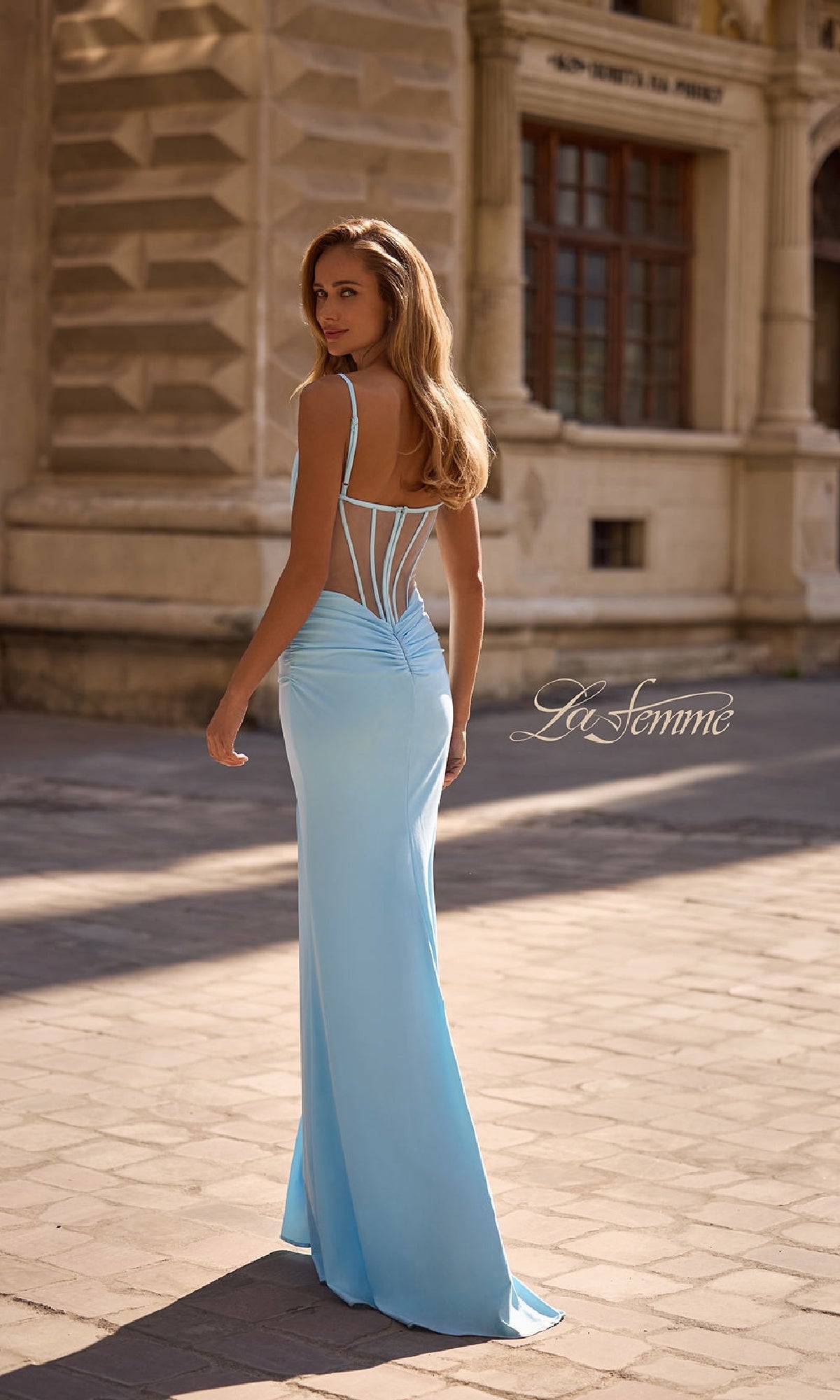 A woman in the Long Jersey Corset Prom Dress: La Femme 32962, featuring a light blue hue and sheer strappy back, stands outdoors on a sunlit stone walkway.